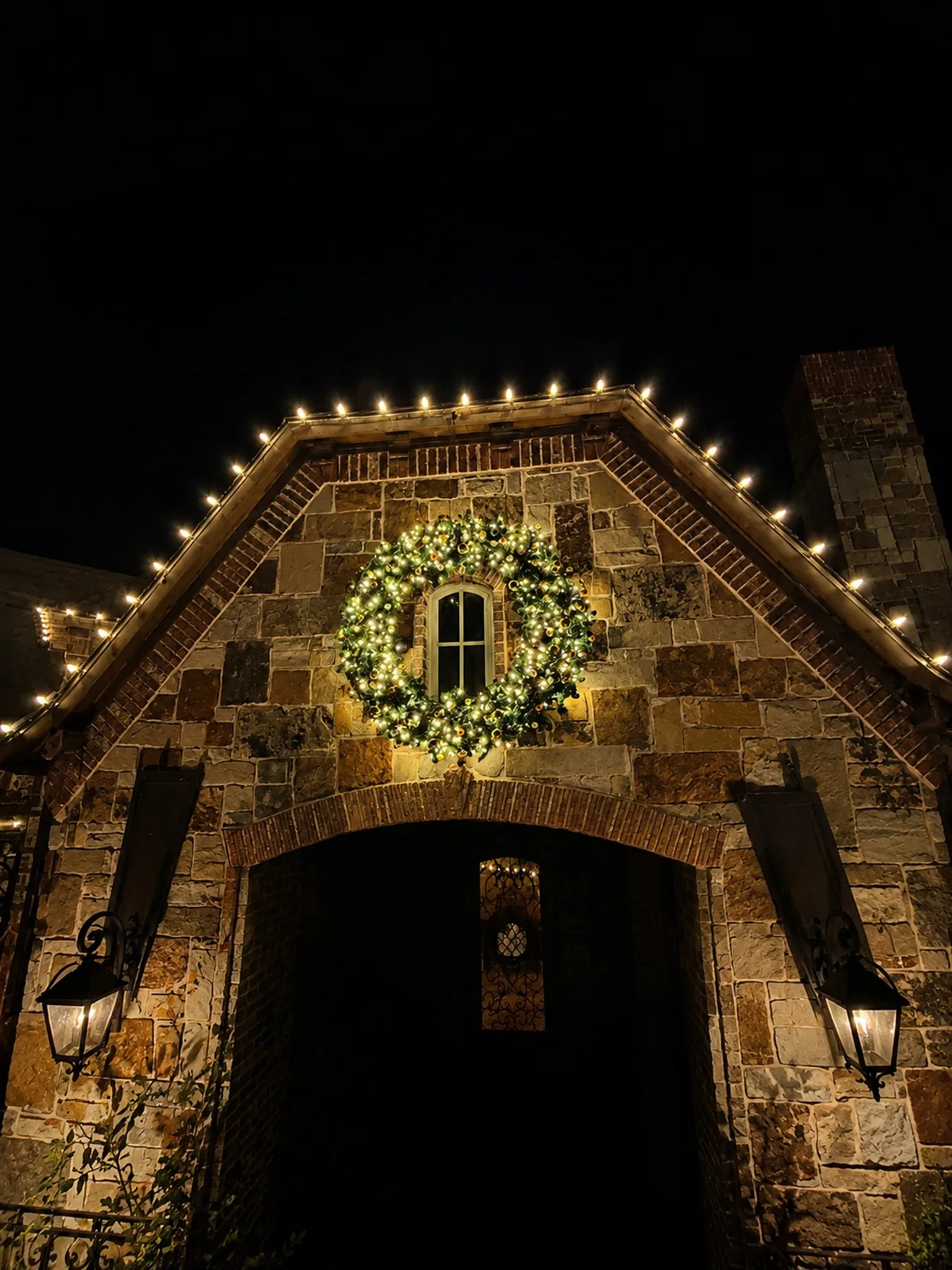 Large illuminated Christmas wreath mounted above a stone arched entry with warm white roofline lights