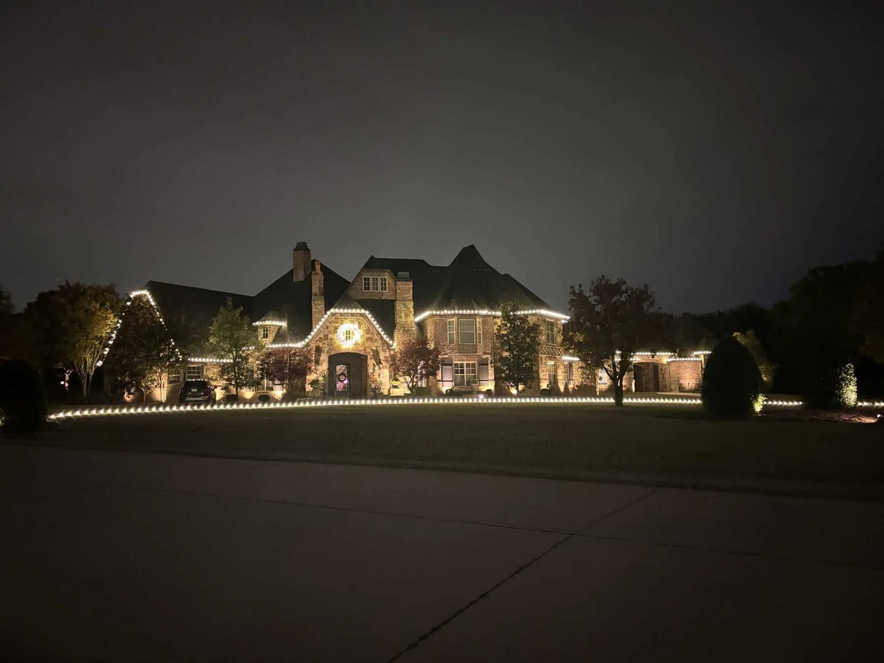 Large estate with warm white roofline, a lit wreath, and lawn edge lighting