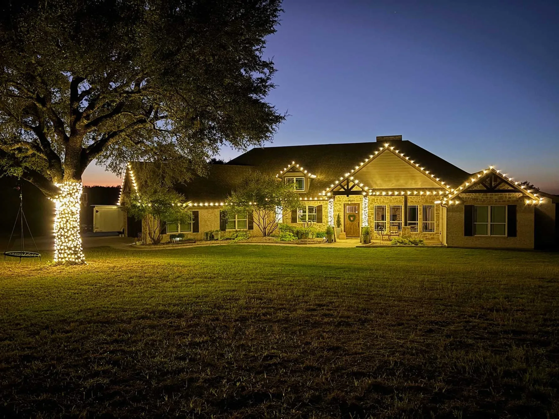 Warm white roofline lighting on a stone home with a wrapped oak tree at dusk