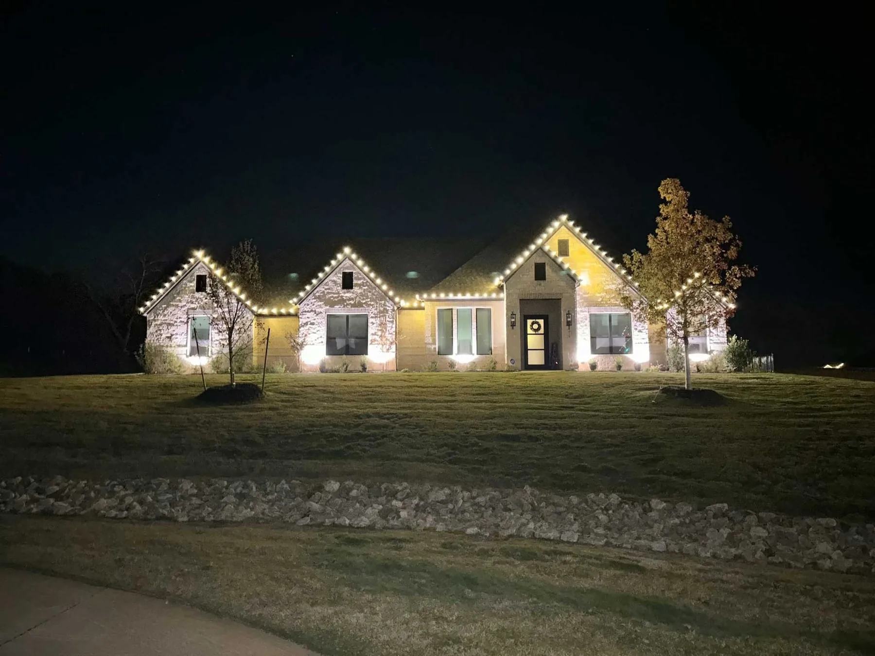 Warm white roofline lighting on a stone home with wrapped trees and a wreath on the front door