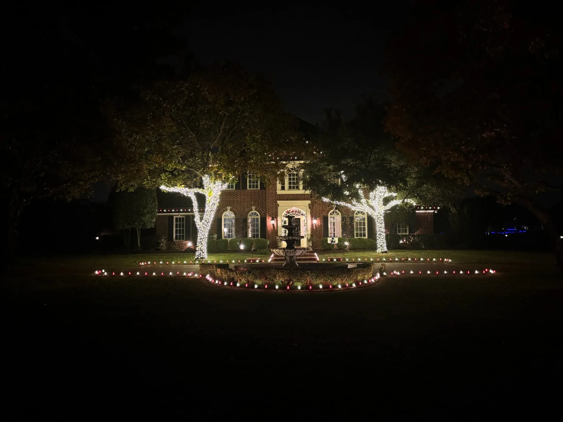Two large trees wrapped in warm white lights beside a lit fountain